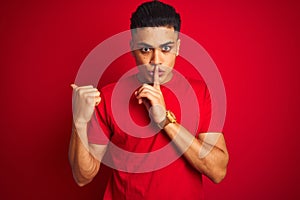 Young brazilian man wearing t-shirt standing over isolated red background asking to be quiet with finger on lips pointing with
