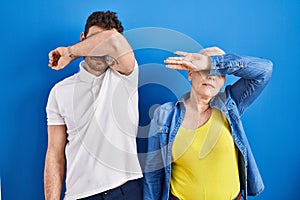 Young brazilian mother and son standing over blue background covering eyes with arm, looking serious and sad