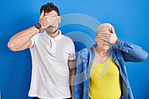 Young brazilian mother and son standing over blue background covering eyes with hand, looking serious and sad