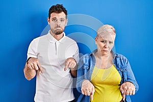 Young brazilian mother and son standing over blue background pointing down looking sad and upset, indicating direction with