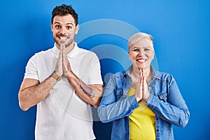 Young brazilian mother and son standing over blue background praying with hands together asking for forgiveness smiling confident