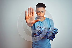 Young brazilian shopkeeper man holding jeans standing over isolated white background with open hand doing stop sign with serious