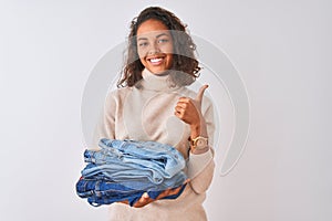 Young brazilian shopkeeper woman holding pile of jeans over isolated white background happy with big smile doing ok sign, thumb up