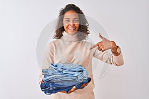 Young brazilian shopkeeper woman holding pile of jeans over isolated white background very happy pointing with hand and finger