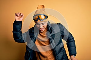 Young brazilian skier man wearing snow sportswear and ski goggles over yellow background Dancing happy and cheerful, smiling