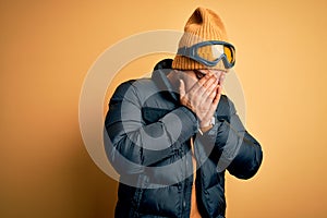 Young brazilian skier man wearing snow sportswear and ski goggles over yellow background with sad expression covering face with
