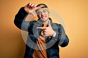 Young brazilian skier man wearing snow sportswear and ski goggles over yellow background smiling making frame with hands and