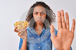 Young brazilian woman holding bowl with macaroni pasta over isolated white background with open hand doing stop sign with serious