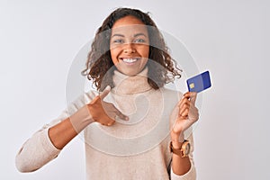 Young brazilian woman holding credit card standing over isolated white background with surprise face pointing finger to himself