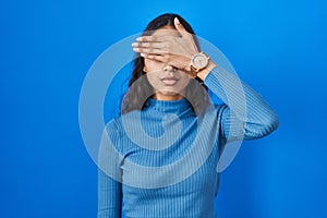 Young brazilian woman standing over blue isolated background covering eyes with hand, looking serious and sad