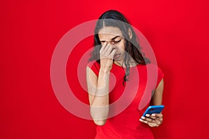 Young brazilian woman using smartphone over red background tired rubbing nose and eyes feeling fatigue and headache