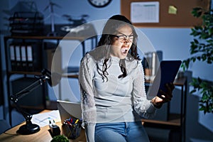 Young brazilian woman using touchpad at night working at the office angry and mad screaming frustrated and furious, shouting with