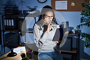 Young brazilian woman using touchpad at night working at the office asking to be quiet with finger on lips
