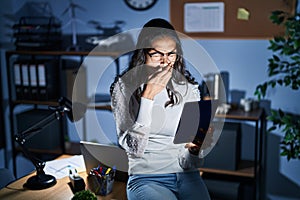 Young brazilian woman using touchpad at night working at the office bored yawning tired covering mouth with hand