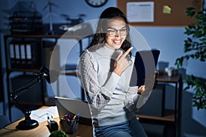 Young brazilian woman using touchpad at night working at the office cheerful with a smile on face pointing with hand and finger up