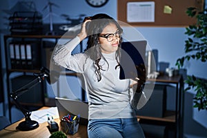 Young brazilian woman using touchpad at night working at the office confuse and wondering about question