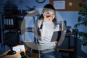 Young brazilian woman using touchpad at night working at the office doing ok gesture with hand smiling, eye looking through