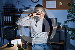 Young brazilian woman using touchpad at night working at the office doing peace symbol with fingers over face, smiling cheerful