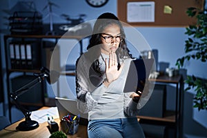 Young brazilian woman using touchpad at night working at the office doing stop sing with palm of the hand