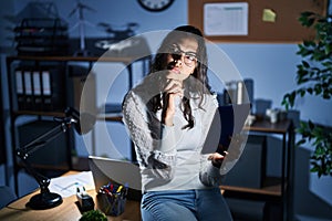 Young brazilian woman using touchpad at night working at the office with hand on chin thinking about question, pensive expression