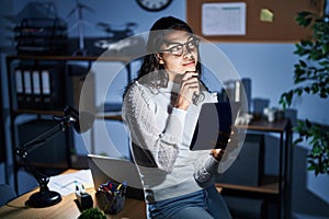 Young brazilian woman using touchpad at night working at the office looking confident at the camera smiling with crossed arms and