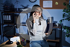 Young brazilian woman using touchpad at night working at the office mouth and lips shut as zip with fingers