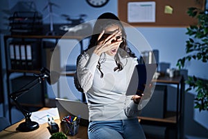 Young brazilian woman using touchpad at night working at the office peeking in shock covering face and eyes with hand, looking