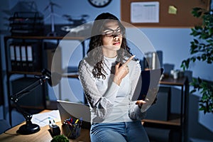 Young brazilian woman using touchpad at night working at the office pointing with hand finger to the side showing advertisement,