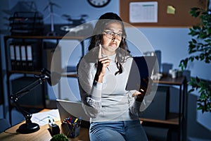 Young brazilian woman using touchpad at night working at the office pointing up looking sad and upset, indicating direction with