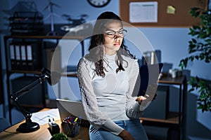 Young brazilian woman using touchpad at night working at the office relaxed with serious expression on face