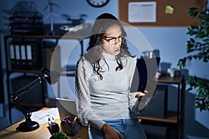 Young brazilian woman using touchpad at night working at the office in shock face, looking skeptical and sarcastic, surprised with