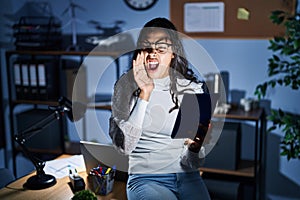 Young brazilian woman using touchpad at night working at the office shouting and screaming loud to side with hand on mouth