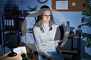 Young brazilian woman using touchpad at night working at the office skeptic and nervous, frowning upset because of problem