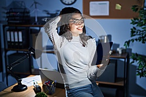 Young brazilian woman using touchpad at night working at the office smiling confident touching hair with hand up gesture, posing