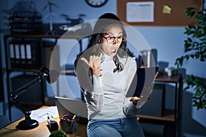 Young brazilian woman using touchpad at night working at the office surprised pointing with hand finger to the side, open mouth