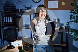 Young brazilian woman using touchpad at night working at the office very happy and excited doing winner gesture with arms raised,