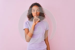 Young brazilian woman wearing t-shirt standing over isolated pink background asking to be quiet with finger on lips