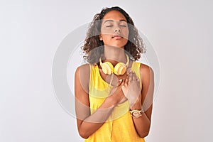 Young brazilian woman wearing yellow headphones over isolated white background smiling with hands on chest with closed eyes and