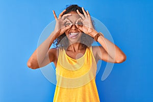 Young brazilian woman wearing yellow t-shirt standing over isolated blue background doing ok gesture like binoculars sticking