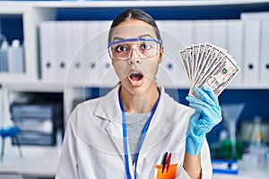 Young brazilian woman working at scientist laboratory holding money scared and amazed with open mouth for surprise, disbelief face