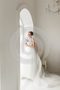 Young bride in a beautiful dress holding a bouquet of flowers posing near window in bright white studio. Wedding concept