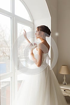 Young bride in a beautiful dress holding a bouquet of flowers posing near window in bright white studio. Wedding concept