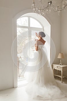 Young bride in a beautiful dress holding a bouquet of flowers posing near window in bright white studio. Wedding concept