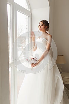 Young bride in a beautiful dress holding a bouquet of flowers posing near window in bright white studio. Wedding concept