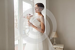 Young bride in a beautiful dress holding a bouquet of flowers posing near window in bright white studio. Wedding concept