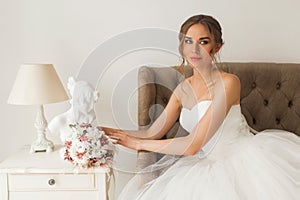 Young bride in a beautiful dress sitting at home with nice white interior. Wedding concept.
