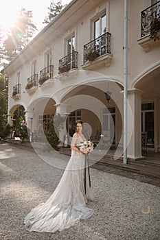 Young bride in fashionable lace wedding dress posing with bridal bouquet outdoors near the villa