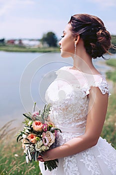 Young bride in a wedding dress is standing on the river bank with a bouquet of flowers
