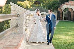 Young bride in white dress and groom walking holding hands near old castle