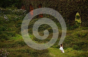 Young bride in white dress running barefoot in field between bushes. Small figure in green fairy tale landscape